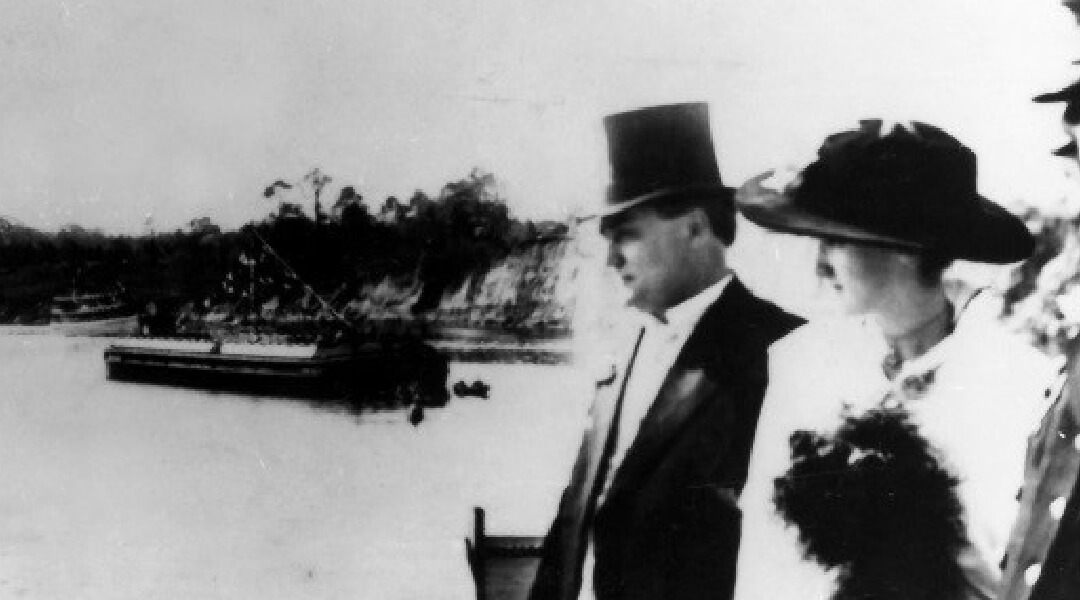 Old photo of well-dressed couple with channel in the background as boat passes.